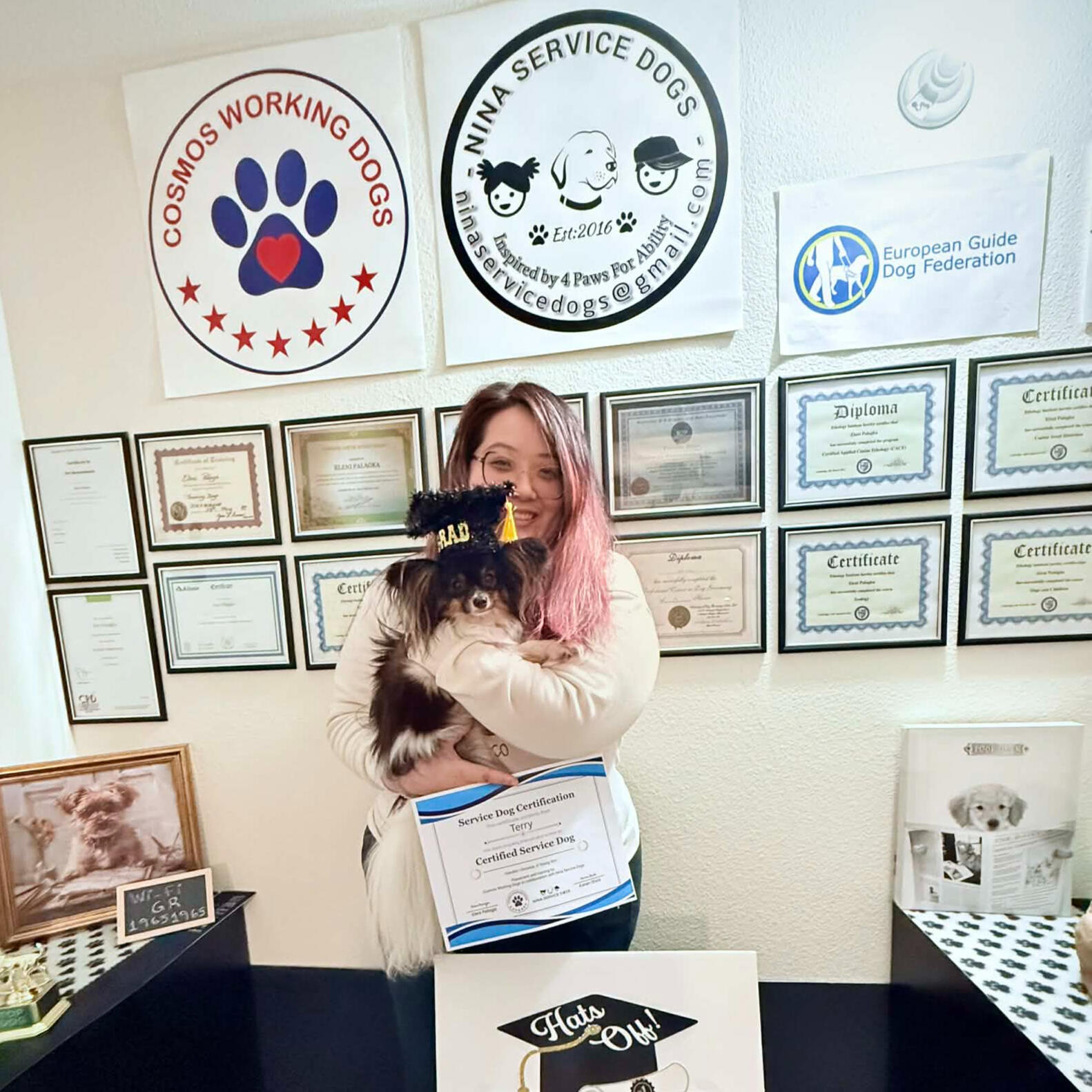 Christine wiseflower Kim holding her Papillon service dog Terry at his certification ceremony, standing in front of a wall of service dog training diplomas.